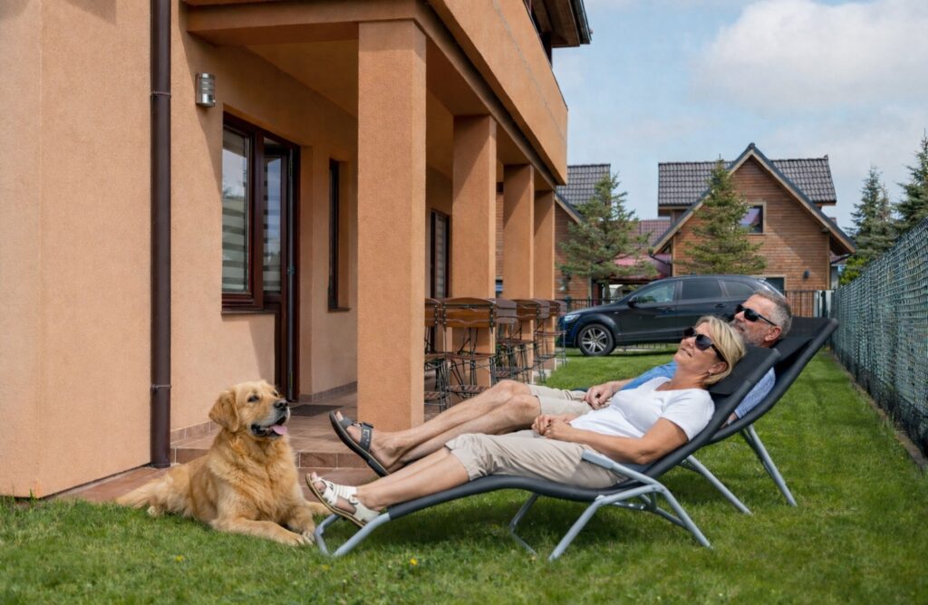 Zwei Hundebesitzer liegen auf Sonnenliegen und der Hund liegt auf dem Rasen vor dem Holiday House Europa in Rewal